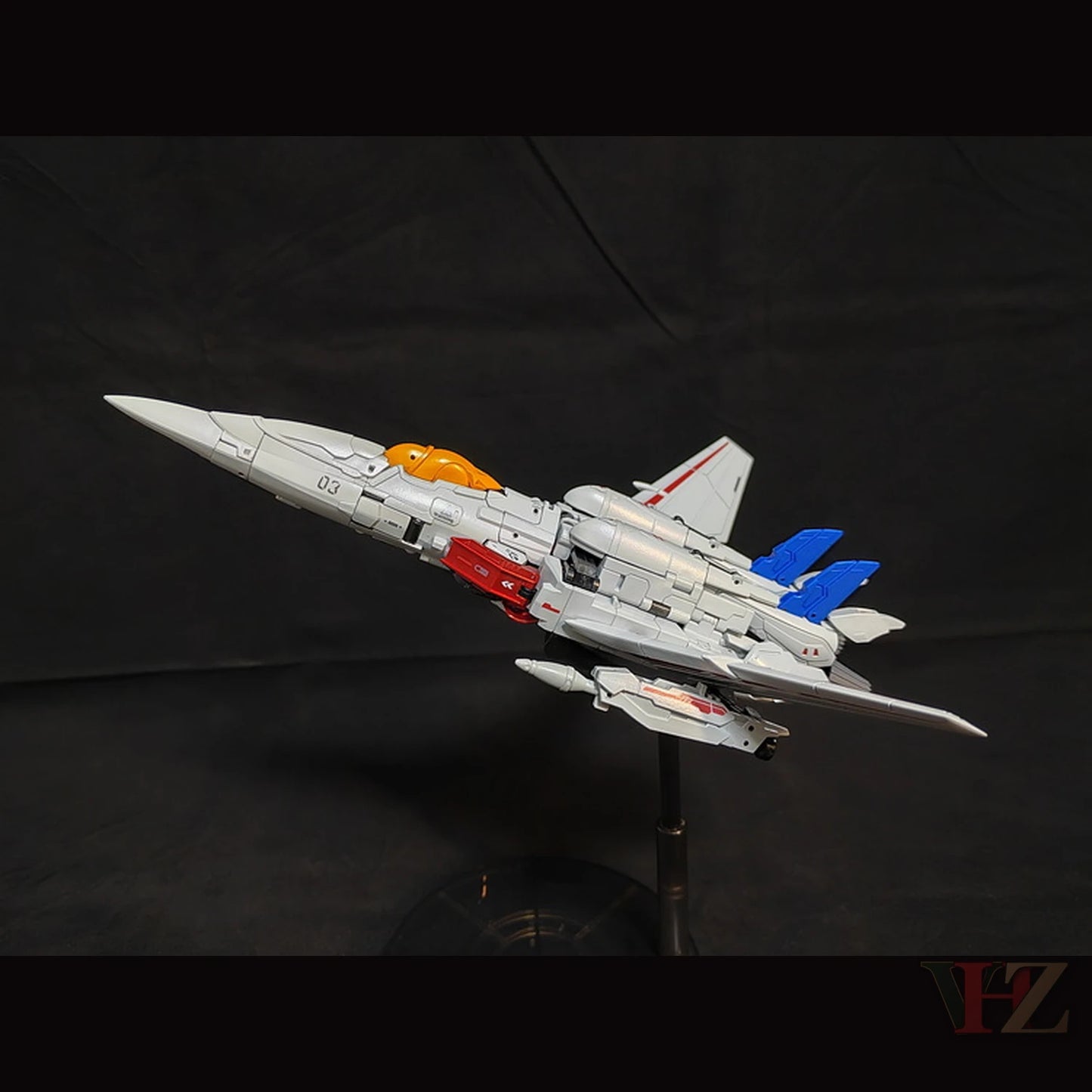 Side view of Collection Space CS-03 Starqueen Starscream jet mode figure, gray with red and blue accents, orange cockpit, "03" marking, on black stand.