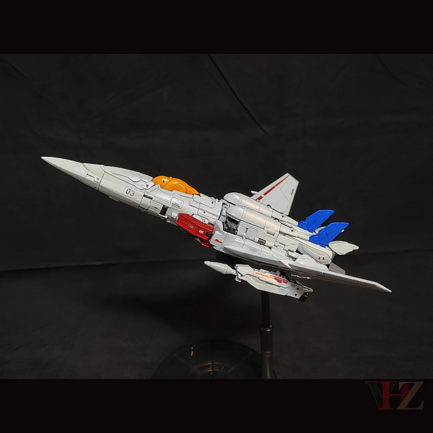 Side view of Collection Space CS-03 Starqueen Starscream jet mode figure, gray with red and blue accents, orange cockpit, "03" marking, on black stand.